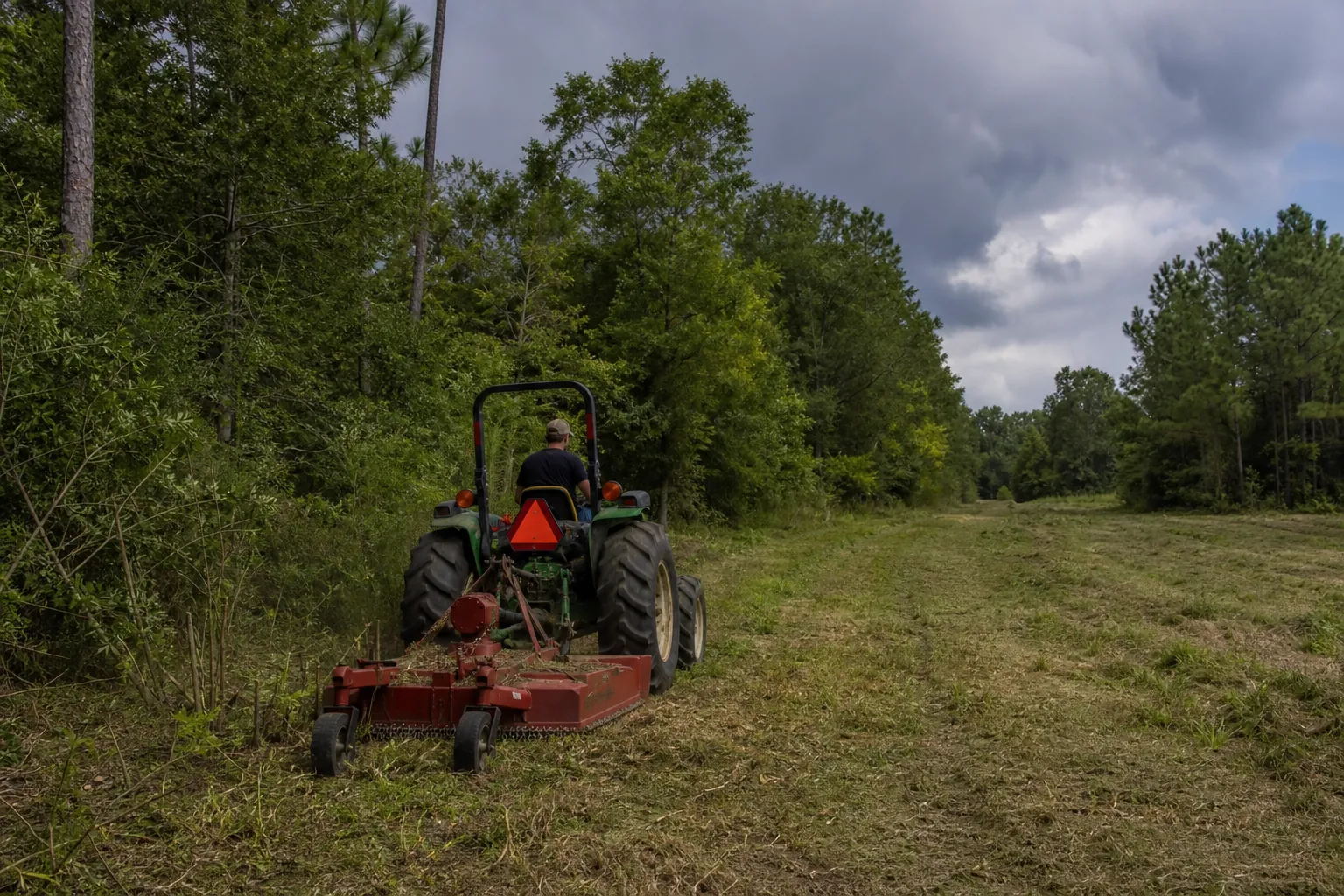 Tractor Brush Hogging a Wooded Clearing A person drives a tractor with a red brush hog along a freshly cut clearing beside dense trees under dark clouds.